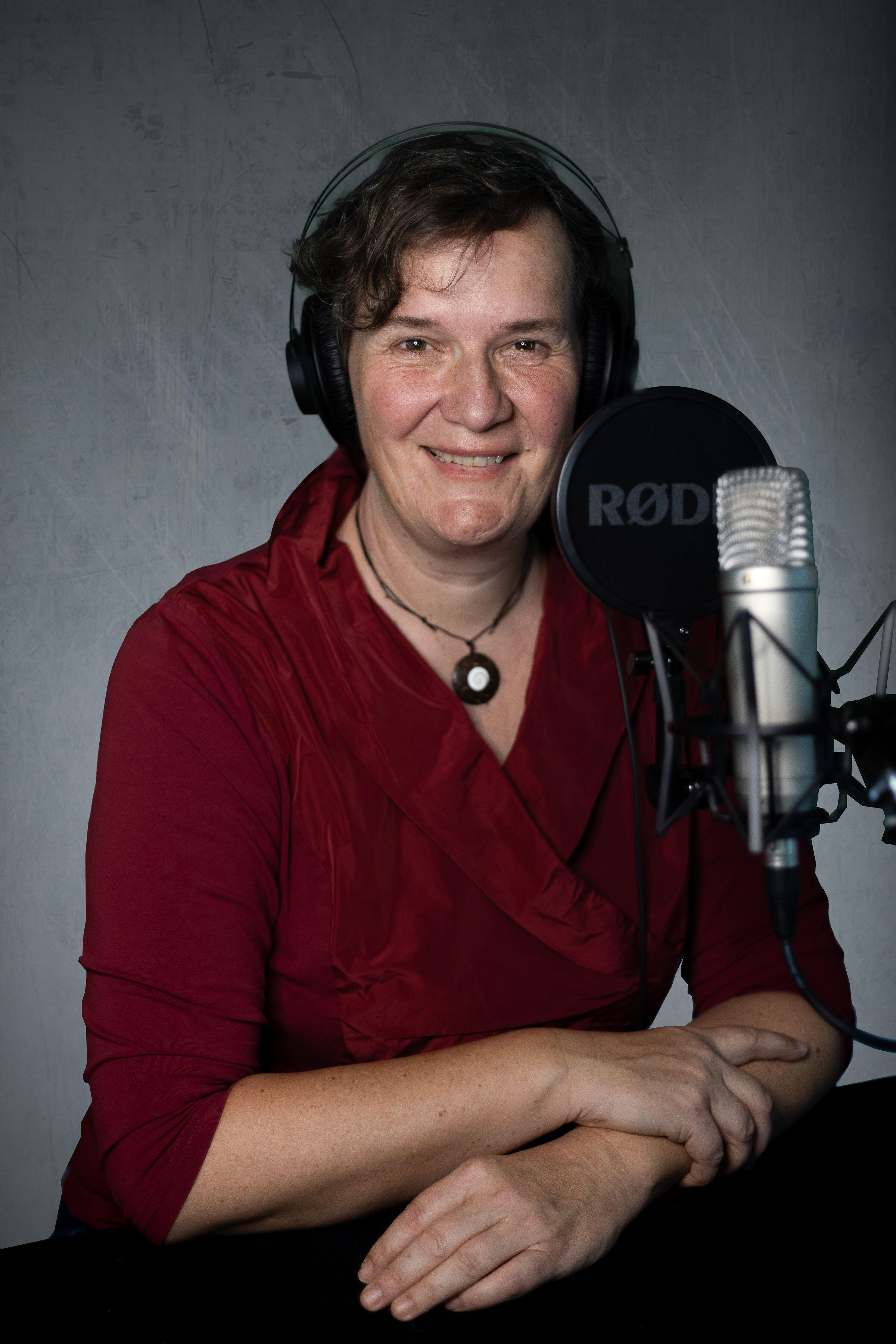 Professionelle Sprecherin Johanna Bahurai #therapeutisches #Storytelling Profilfoto von Johanna Bahurai in roter Bluse mit Kopfhörer auf dem Kopf vor einem Studio-Mikrofon, in die Kamera lächelnd.